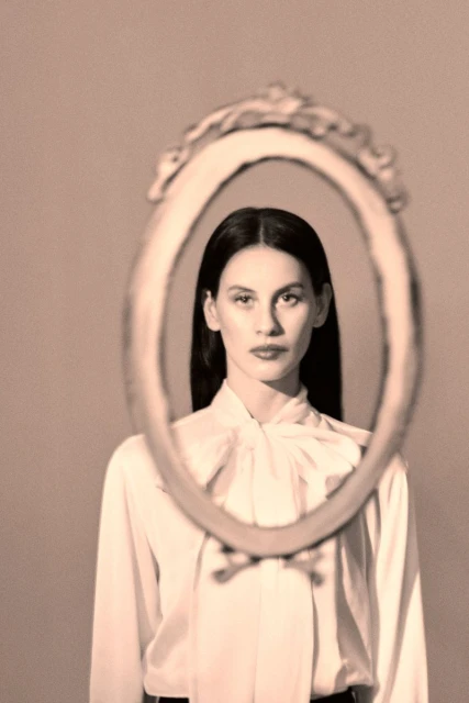 Indoor portrait of a young adult woman looking through an antique mirror in a studio, posing in a vintage fashion style.