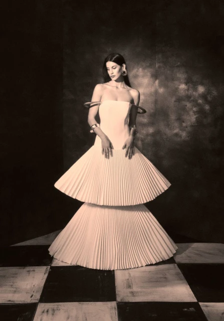A woman poses in a pleated gown against a dark background with a black-and-white checkered floor for a fashion editorial.