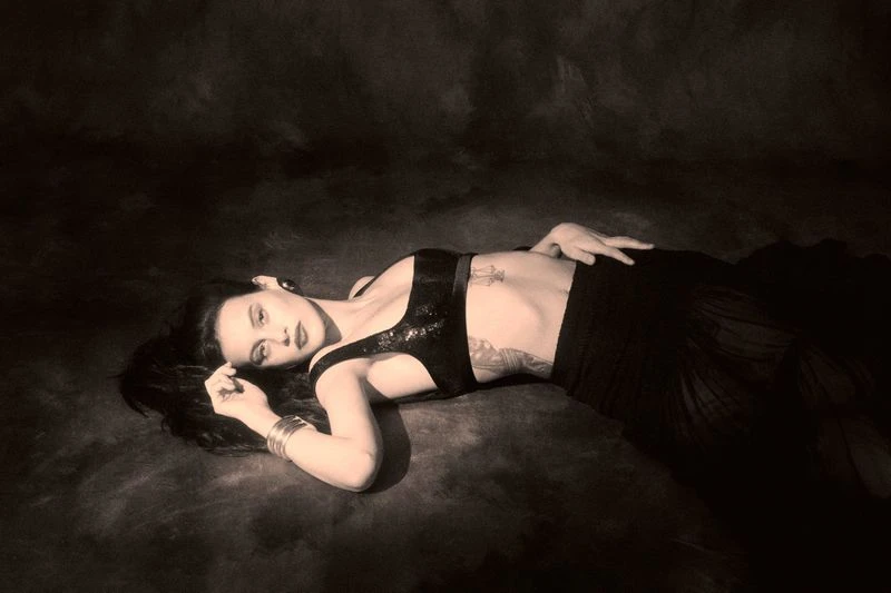 An adult female laying down wearing a top and skirt in a studio with jewelry, make-up, and tattoos.  Monochromatic and moody.