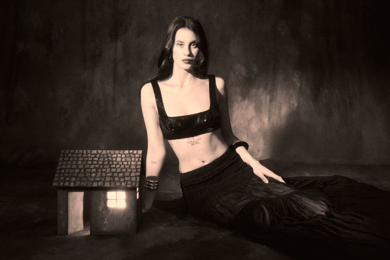Woman poses in a black top and skirt beside a small house with a lit window against a muted background in an indoor setting.