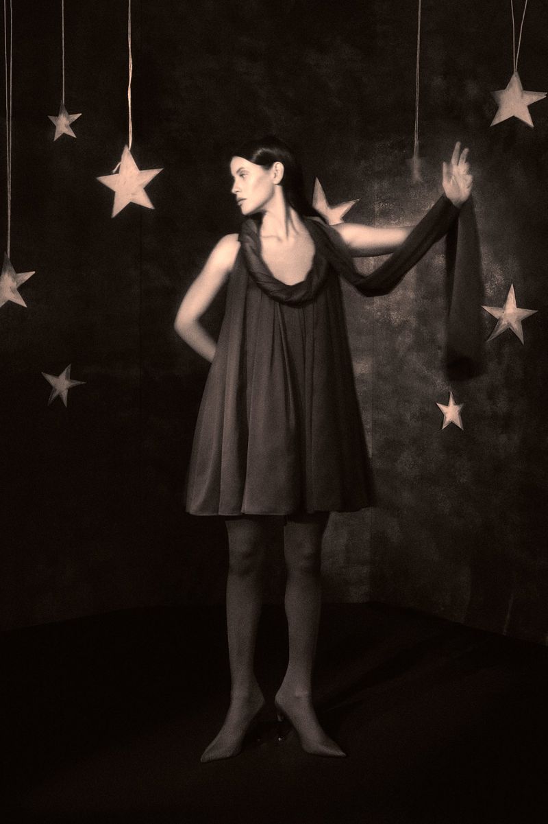 Woman in vintage dress poses in a dark studio surrounded by hanging stars. Sepia-toned, surreal fashion photography with a celestial theme.