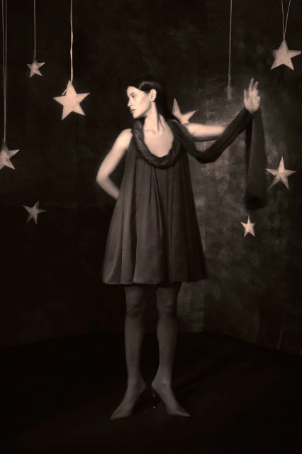 Woman in vintage dress poses in a dark studio surrounded by hanging stars. Sepia-toned, surreal fashion photography with a celestial theme.