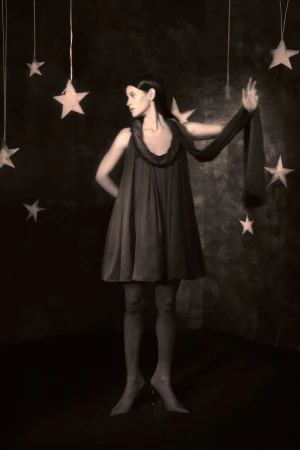 Woman in vintage dress poses in a dark studio surrounded by hanging stars. Sepia-toned, surreal fashion photography with a celestial theme.