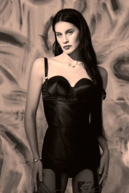 A woman with long, dark hair wearing vintage-style lingerie, posing against a backdrop of abstract painting in monochrome. 