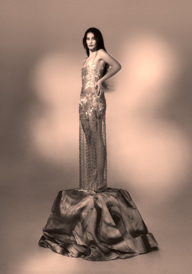 A woman stands on a draped pedestal, wearing a floor-length beaded gown, looking at the camera in a sepia-toned studio shot.