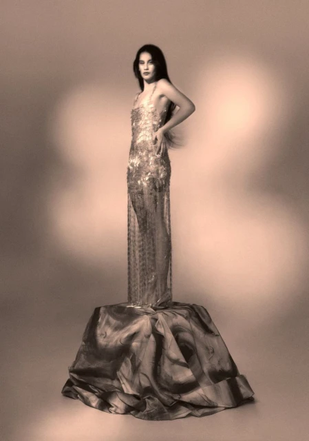 A woman stands on a draped pedestal, wearing a floor-length beaded gown, looking at the camera in a sepia-toned studio shot.