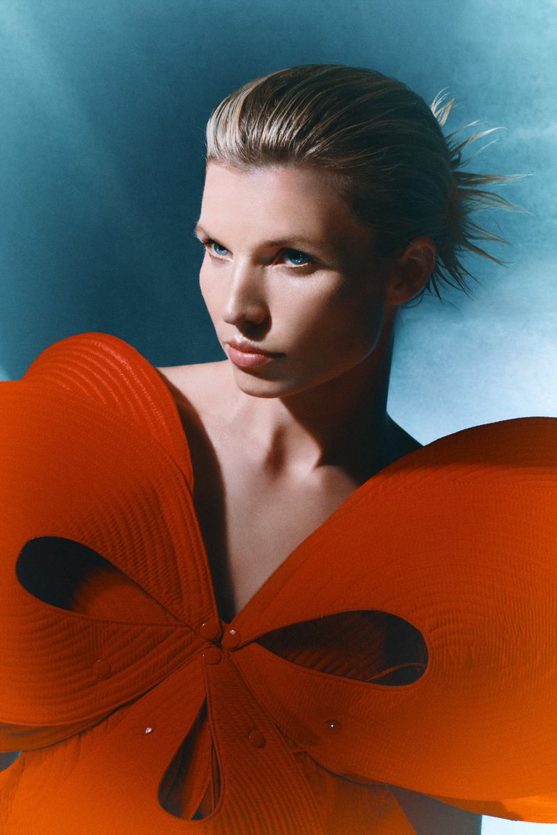 Glamorous fashion portrait of a blonde woman with a unique orange bow-shaped dress against a blue background in a studio setting.