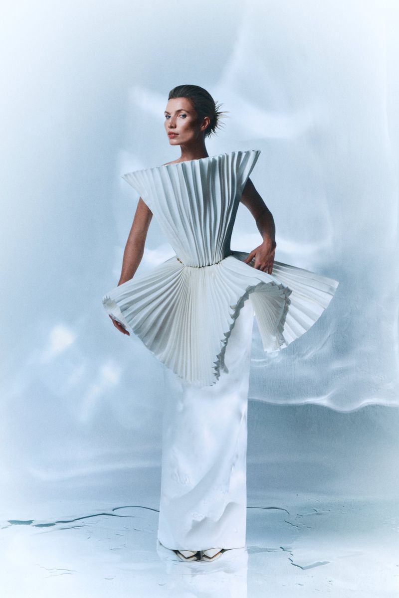 Woman in an elegant white gown with pleated ruffles posing in a studio setting with soft lighting and a reflective floor.