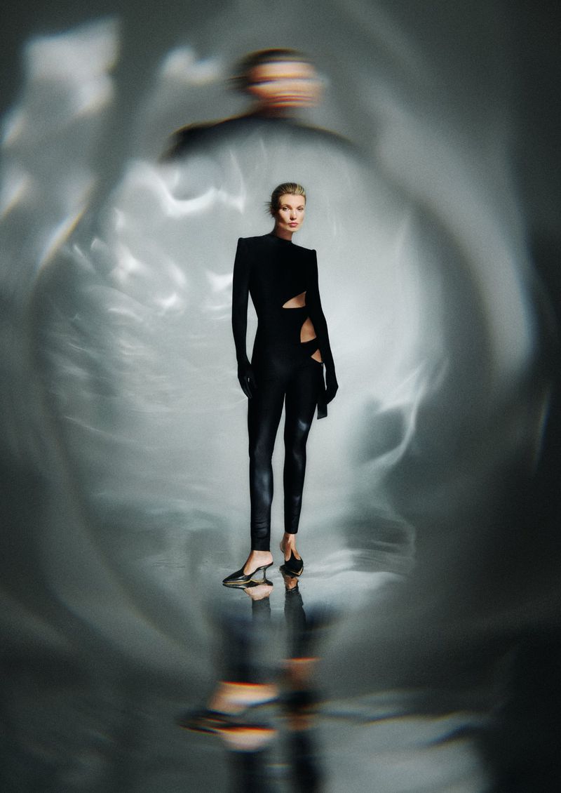 Blond woman in a black cut-out catsuit poses in front of a blurred light background in a studio fashion shot.