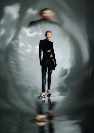 Blond woman in a black cut-out catsuit poses in front of a blurred light background in a studio fashion shot.