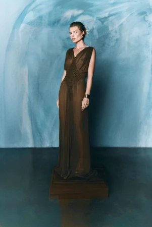 A slender woman in a floor-length sheer dress stands on a wooden platform, posing in front of an abstract blue painted backdrop.