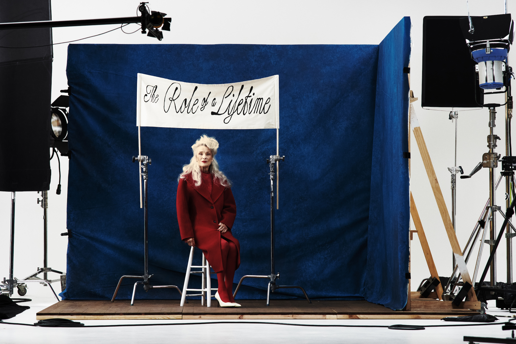 Elegant senior woman in red suit poses on white stool against blue backdrop with 'The Role of a Lifetime' banner in studio setting.