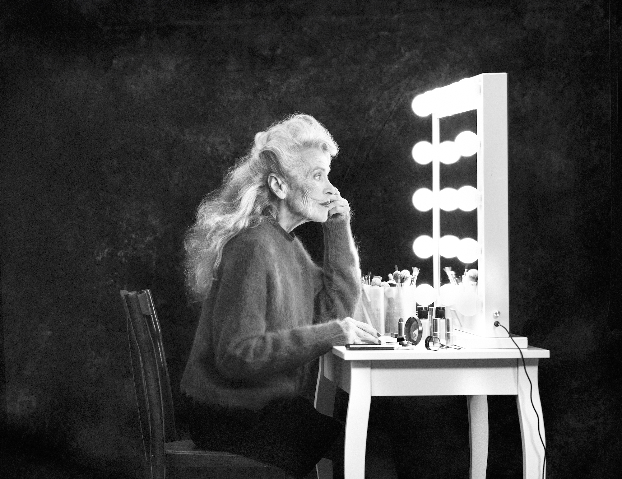 Monochrome portrait of an elderly woman at a vanity, applying makeup, with a light-up mirror and various cosmetics visible on the table.