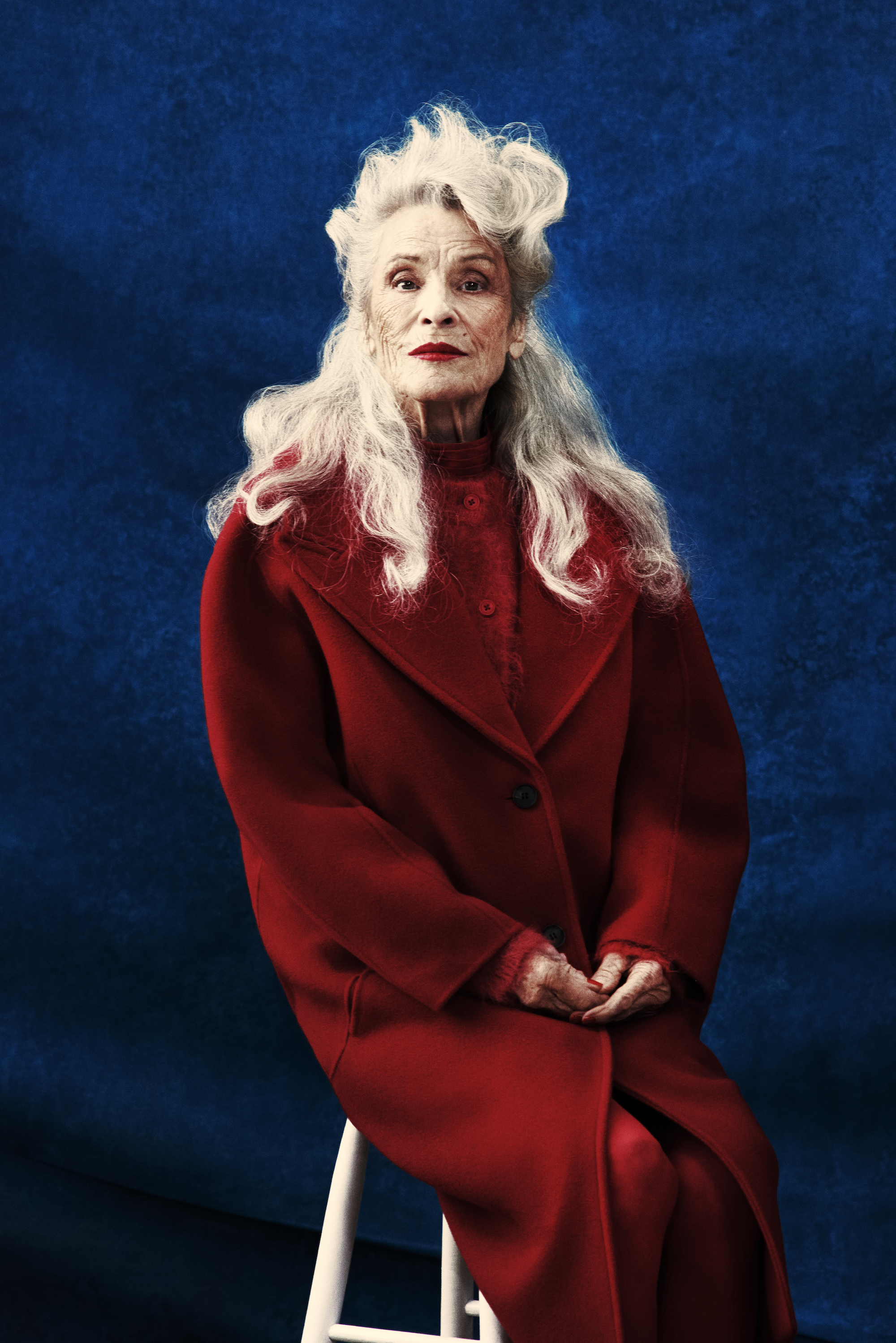 Elderly woman in red coat sitting on stool against a blue background. She has silver hair and red lipstick for a classic portrait.