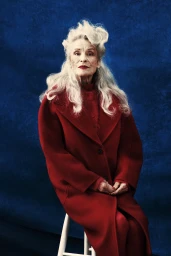 Elderly woman with voluminous white hair wearing a red coat seated on a stool against a textured blue backdrop