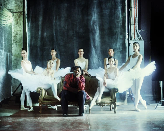 Seven ballet dancers pose on and around a tufted green velvet sofa in a photo studio with gray walls and a backdrop in dim light.
