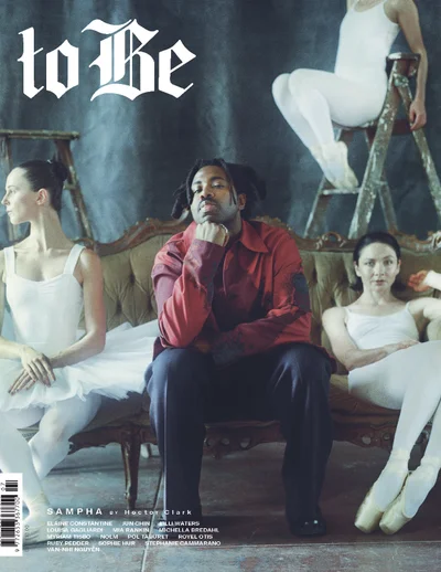 Musician Sampha seated on a vintage sofa flanked by three dancers in white leotards and pointe shoes in an editorial portrait.