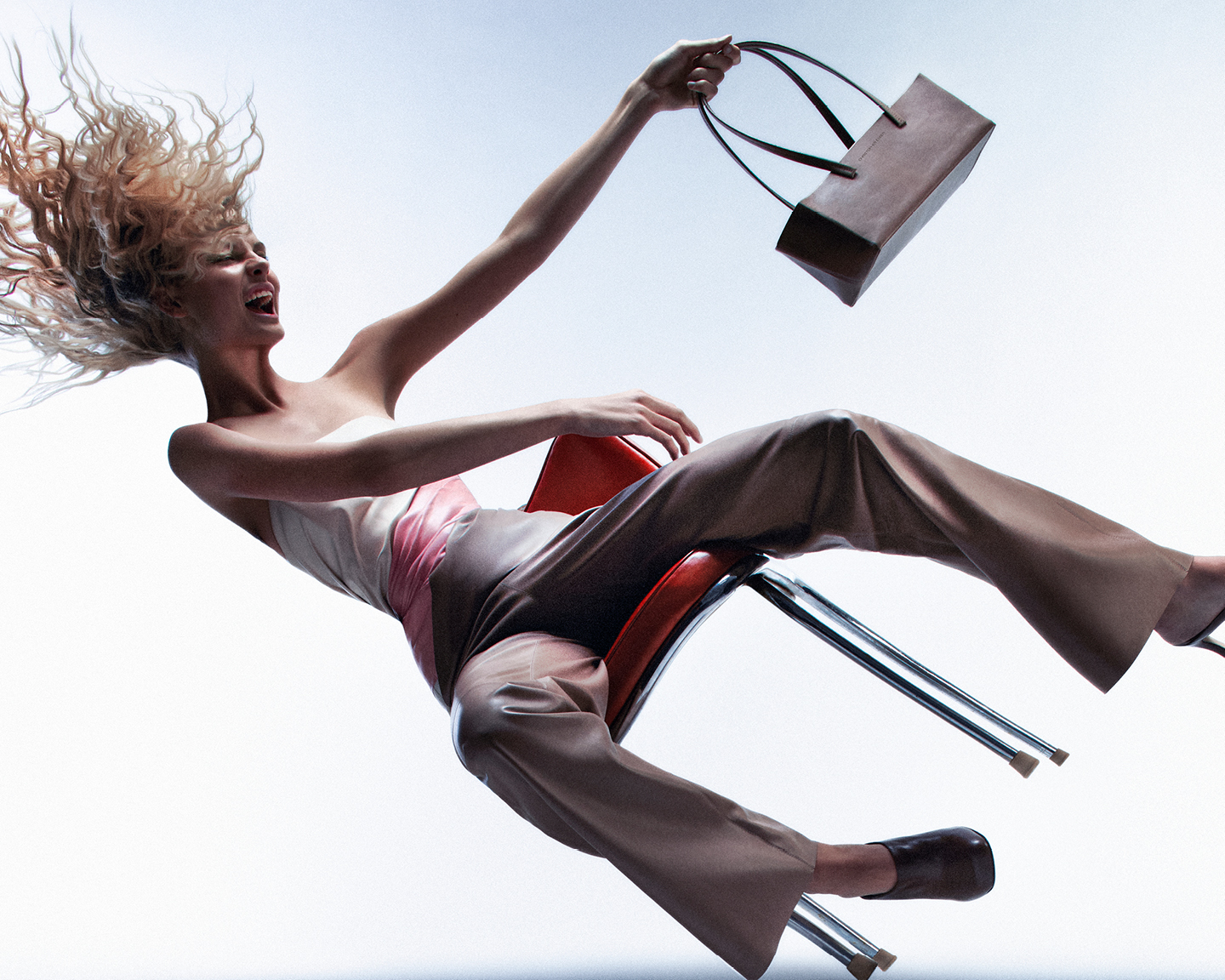 Fashion shot of a woman falling from a chair, holding a brown handbag against a bright studio backdrop with flowing blonde hair.