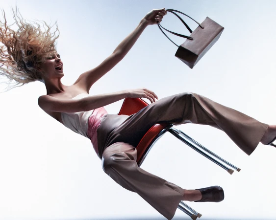 Fashion shot of a woman falling from a chair, holding a brown handbag against a bright studio backdrop with flowing blonde hair.