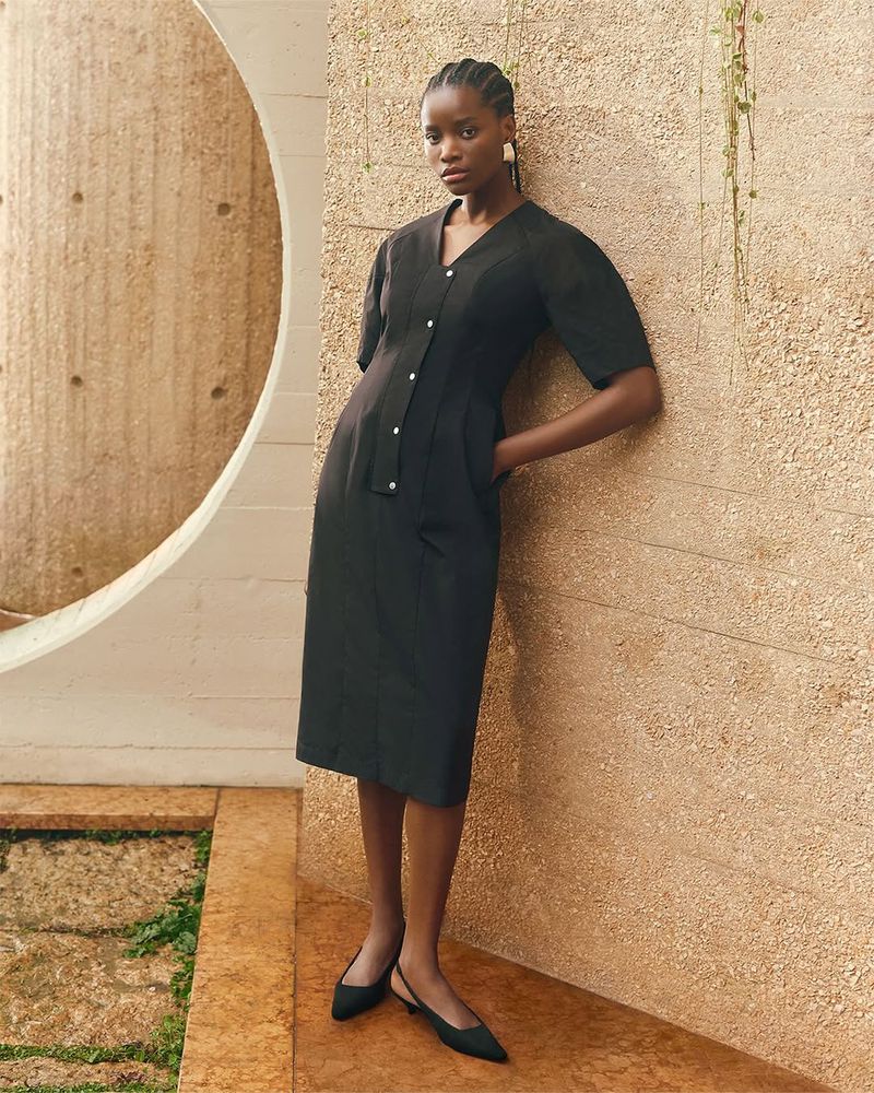 A slender woman in a black midi dress with button-up detail poses against a textured beige wall in a fashion-editorial style indoor shot.