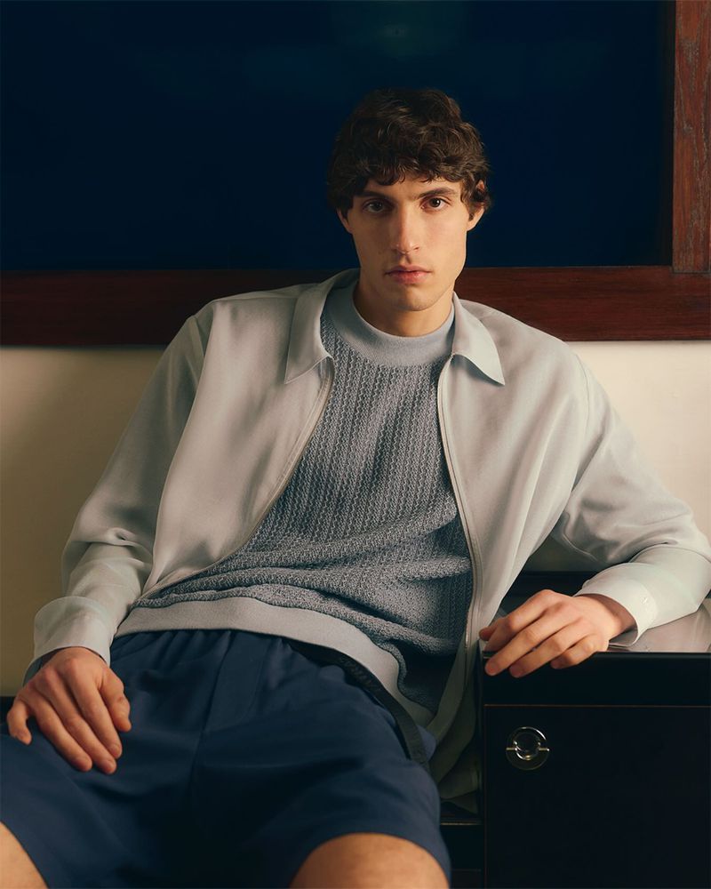 Young man in pale blue jacket and knit sweater poses indoors near a dark chest, bathed in soft, neutral lighting.