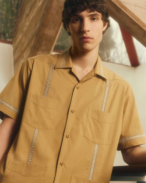 A young adult male poses in a tan, short-sleeved Cuban-collar shirt with decorative embroidery and four pockets against a studio backdrop.