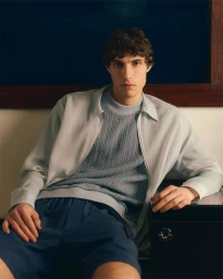 A man in a light blue textured sweater and dark trousers seated indoors for TATRAS Spring-Summer 25