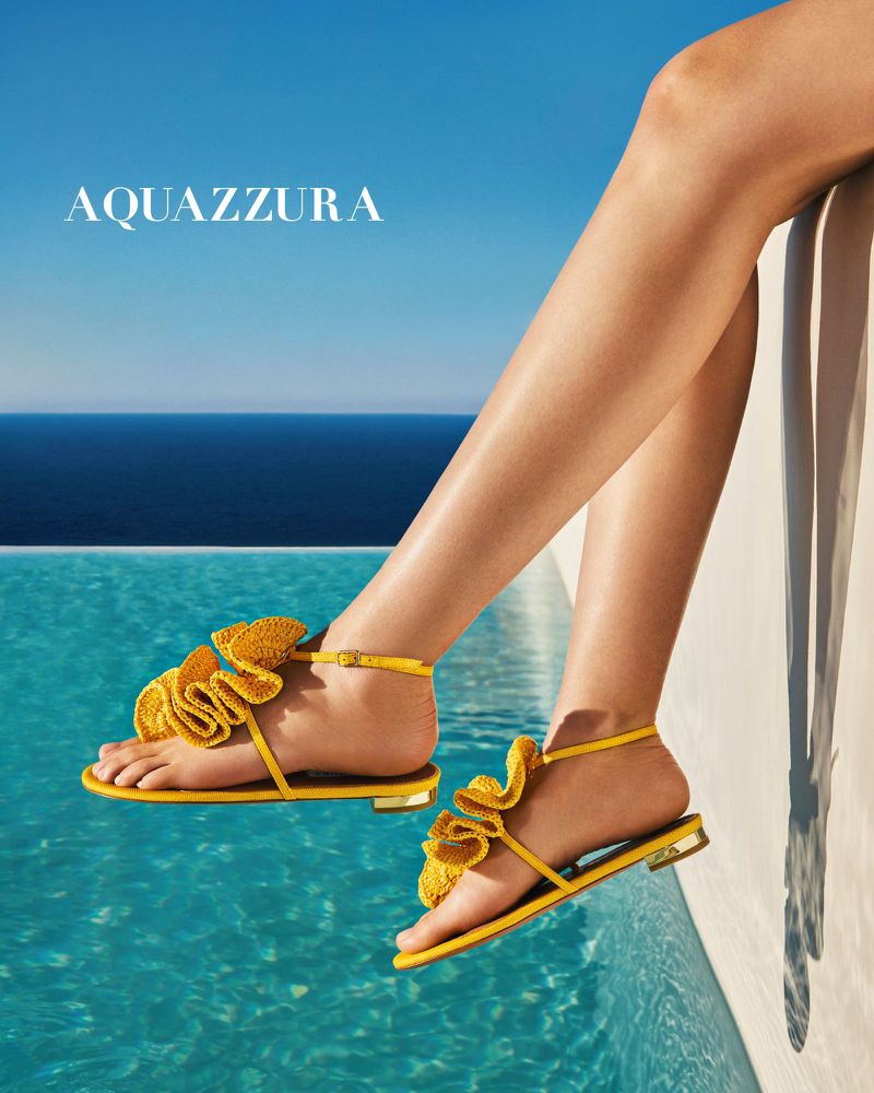 Elevated view of a woman's legs and feet adorned with Aquazzura yellow sandals, against a blue pool and sky backdrop.