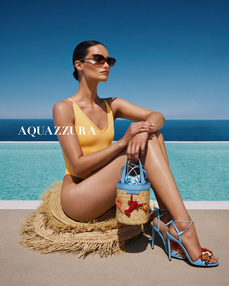 Woman in yellow swimsuit poses with straw purse and heels near a pool with a clear blue sky in a fashion advertisement.