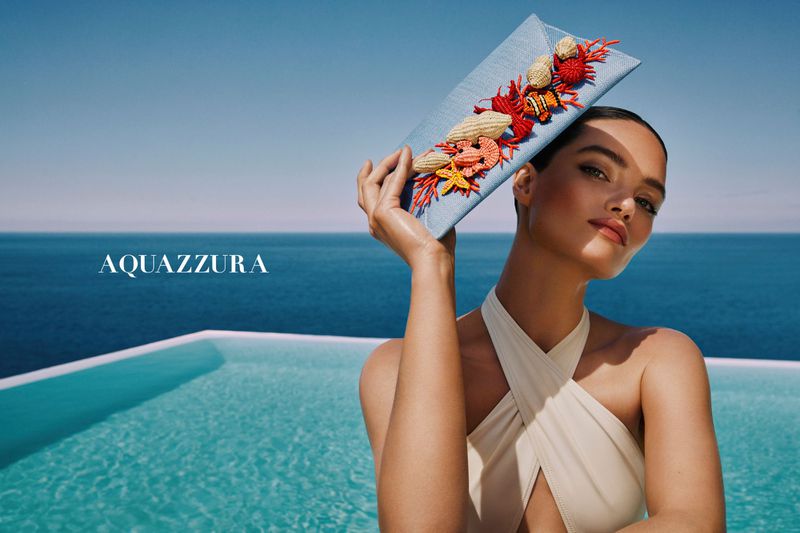 Woman poses with a light blue handbag adorned with red and white shells, starfish, and a clownfish, in front of a pool and ocean.