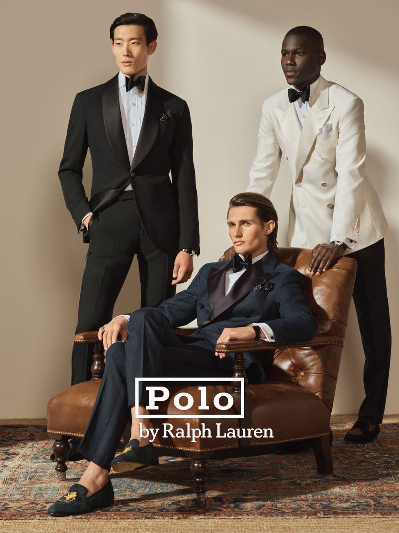 Three men in formalwear pose indoors on and around a leather chair. One man sits, one stands to the left, one leans on chair back.