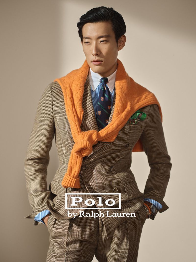 Man in a brown plaid suit with an orange sweater draped over his shoulders, standing in a neutral setting for a fashion editorial.