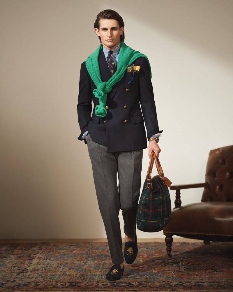 Man in a navy double-breasted blazer, grey trousers, and black loafers, carrying a plaid duffel bag, standing in an indoor setting.