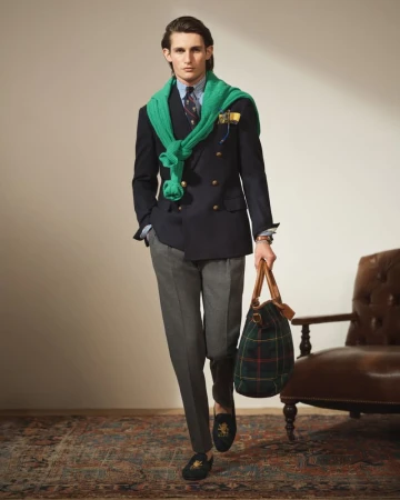 Man in a navy double-breasted blazer, grey trousers, and black loafers, carrying a plaid duffel bag, standing in an indoor setting.
