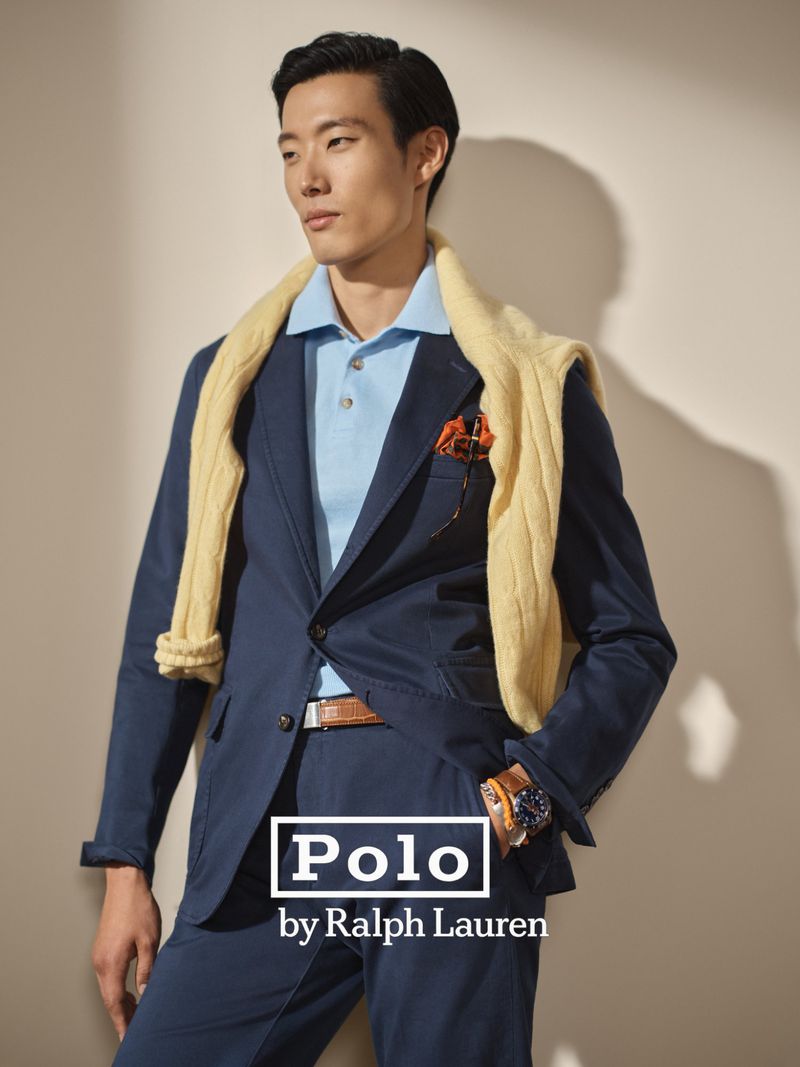 Asian man in a blue suit and light blue polo shirt with a yellow sweater draped around his shoulders posing in a studio.