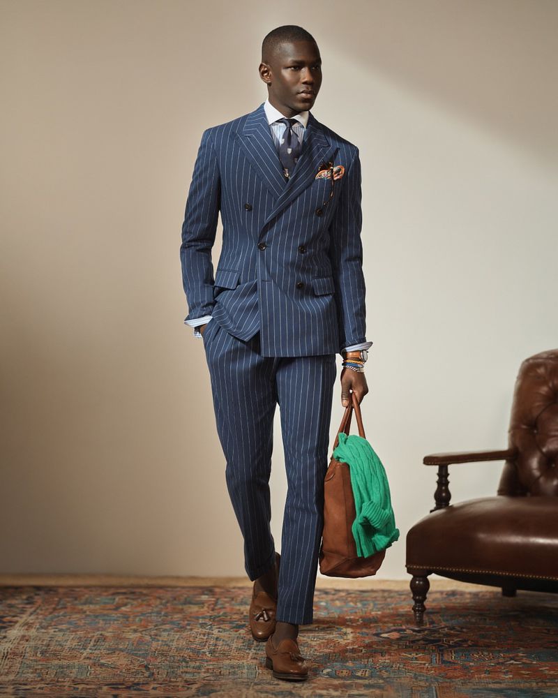 A stylish Black man walks in a pinstripe suit, holding a brown leather bag and wearing brown dress shoes on an ornate rug with a chair.