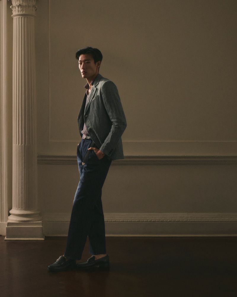 A stylish Asian man poses indoors wearing a blazer, patterned shirt, dark wash denim jeans, and loafers in a fashion portrait.