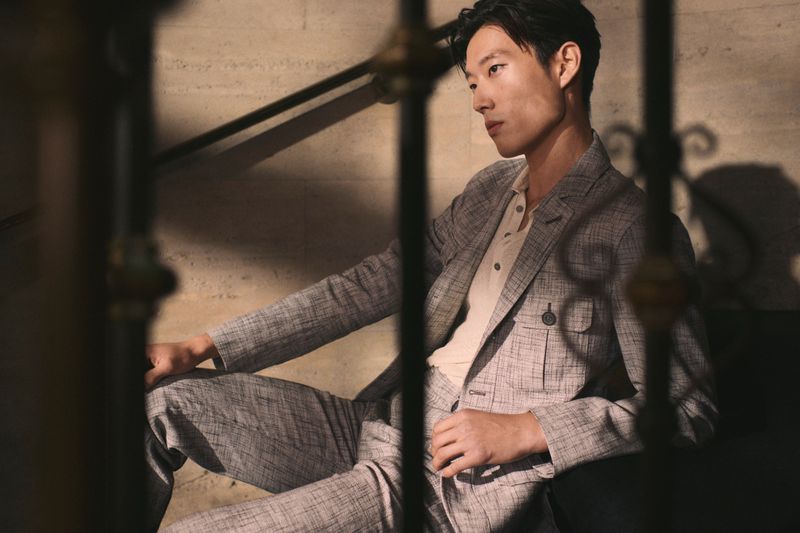 Asian man in a grey suit poses in a dimly lit indoor setting. He is seated, with dark hair, wearing a beige collared shirt.