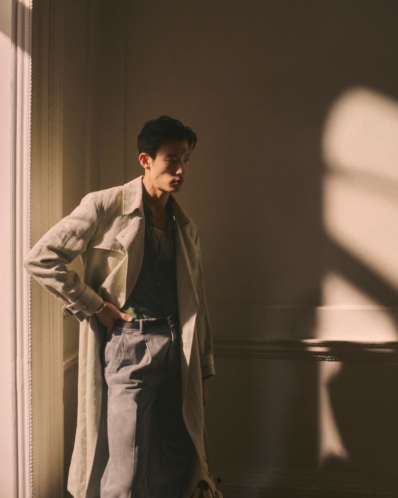 Man in trench coat poses indoors in shadows, wearing a tank top and gray pants, with light entering from a nearby window.