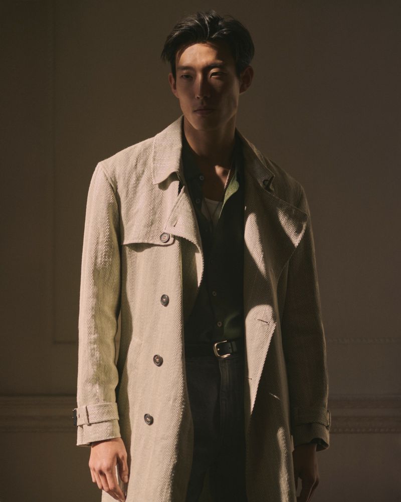 An Asian adult male in a trench coat poses indoors in diffused, warm lighting, showcasing fashion and style.