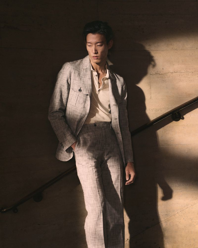 Man poses in suit against light and shadow. He wears a light-colored suit with a matching polo shirt, hand in his pocket.
