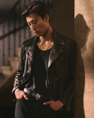 Portrait of Asian man in black leather fringe jacket leaning against wall looking down in medium shot, shallow depth of field, warm lighting.