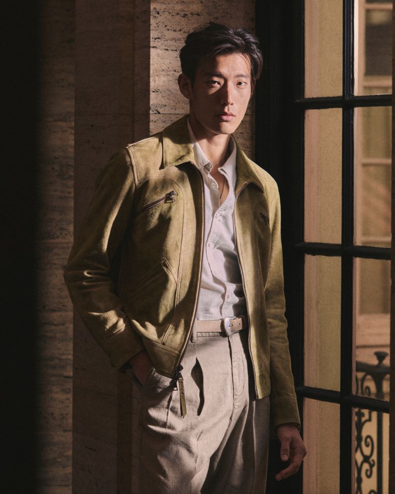Portrait of an Asian male model in a light brown jacket, posing indoors near a window with dark panes. He is wearing a white shirt.