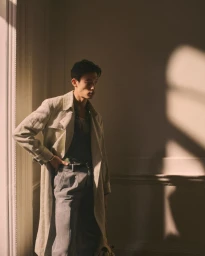 Model wearing a light trench coat and grey trousers posed against a neutral wall indoors with dramatic shadows.