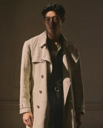 Man wearing a textured beige trench coat over a dark shirt against a neutral wall with dramatic directional lighting.