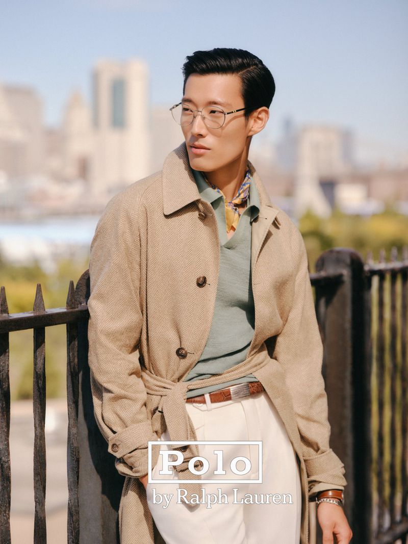 Man in trench coat and glasses poses outdoors against a city backdrop in a fashion shot by Polo Ralph Lauren.