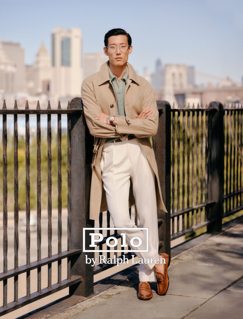 Man in glasses and tan trench coat leans against fence with NYC buildings in the background, wearing Polo by Ralph Lauren attire.