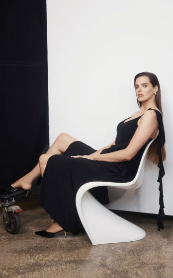 Adult woman in a black dress sits in a white chair in a photo studio with a black and white background, side lighting, and medium shot.