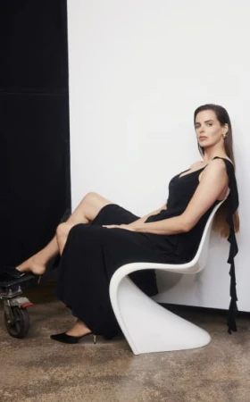 Adult woman in a black dress sits in a white chair in a photo studio with a black and white background, side lighting, and medium shot.