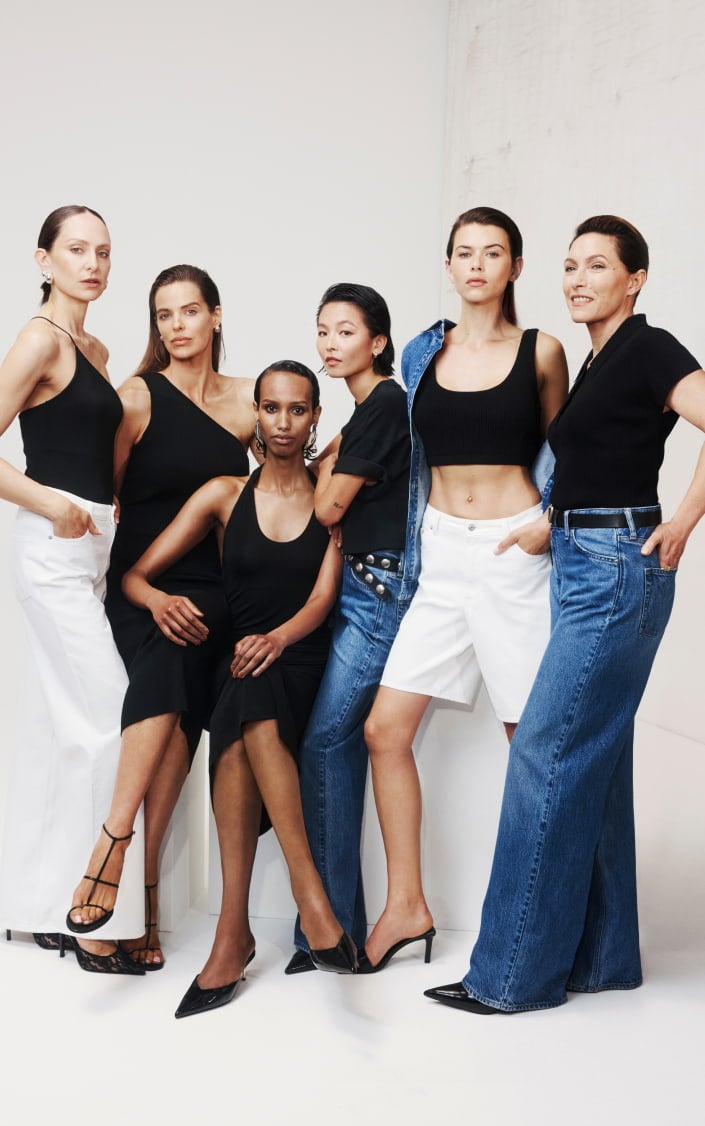 Group of six diverse female models in black tops and jeans or white trousers posing together in a studio setting.